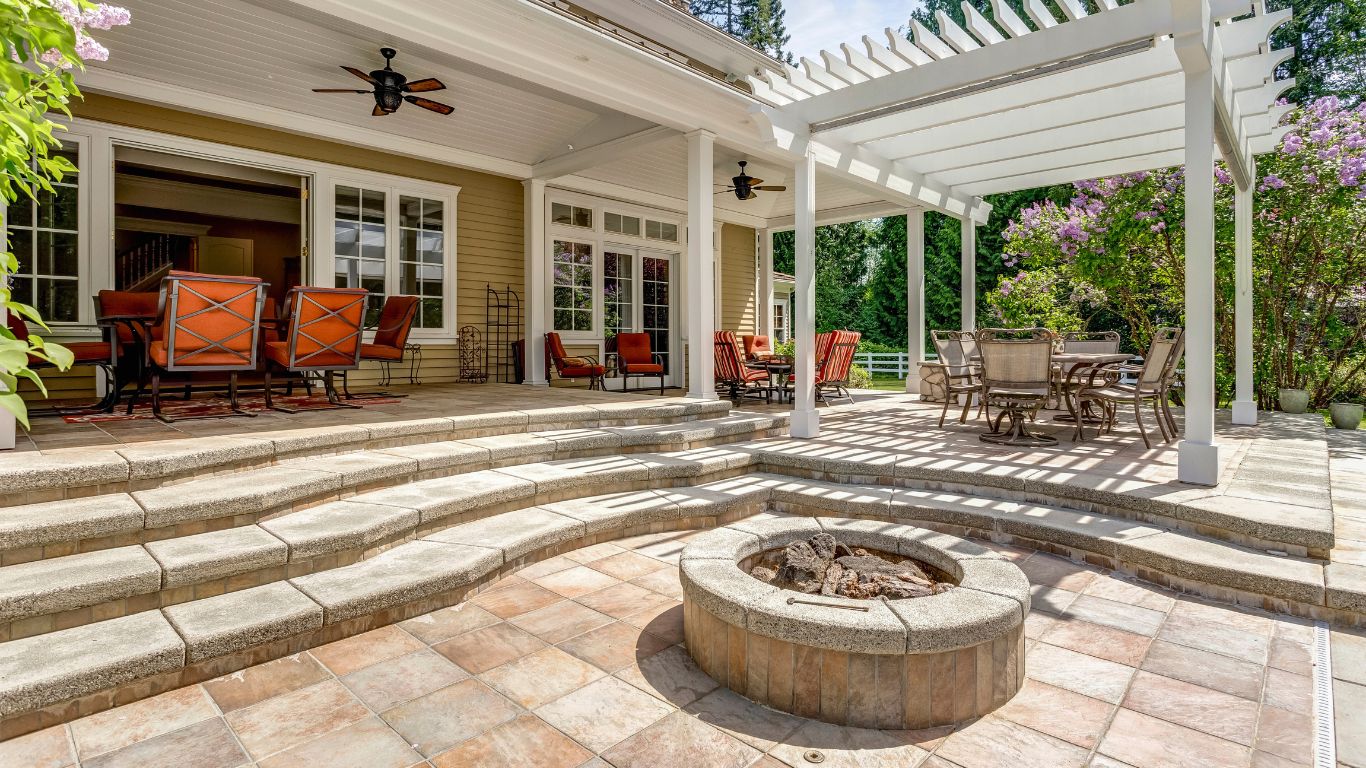Beautiful backyard retreat featuring elegant kitchen pavers, a multi-level patio with cozy outdoor seating, a stylish firepit, and a white pergola-covered dining area surrounded by lush greenery.